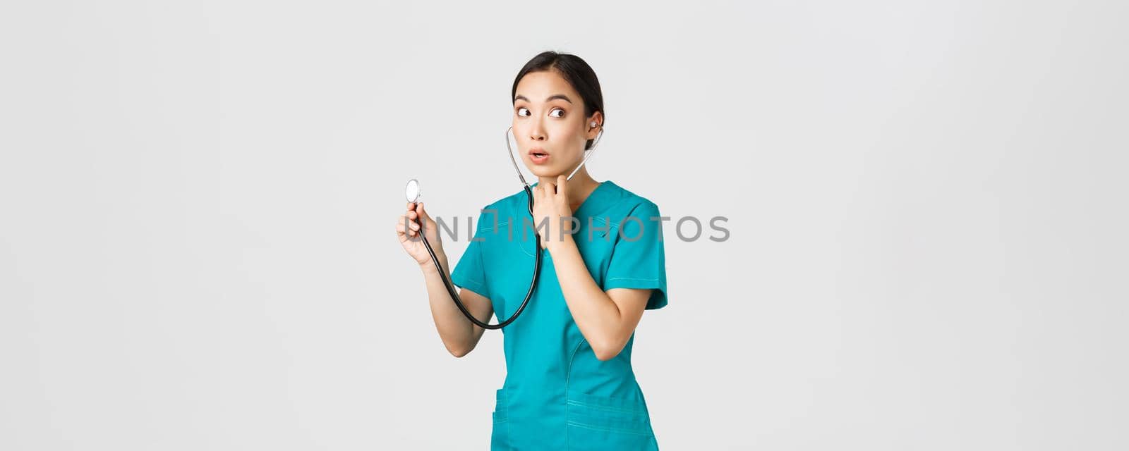 Covid-19, healthcare workers and preventing virus concept. Intrigued asian female doctor, physician in scrubs, looking away thoughtful, listening to patient lungs with stethoscope, white background by Benzoix