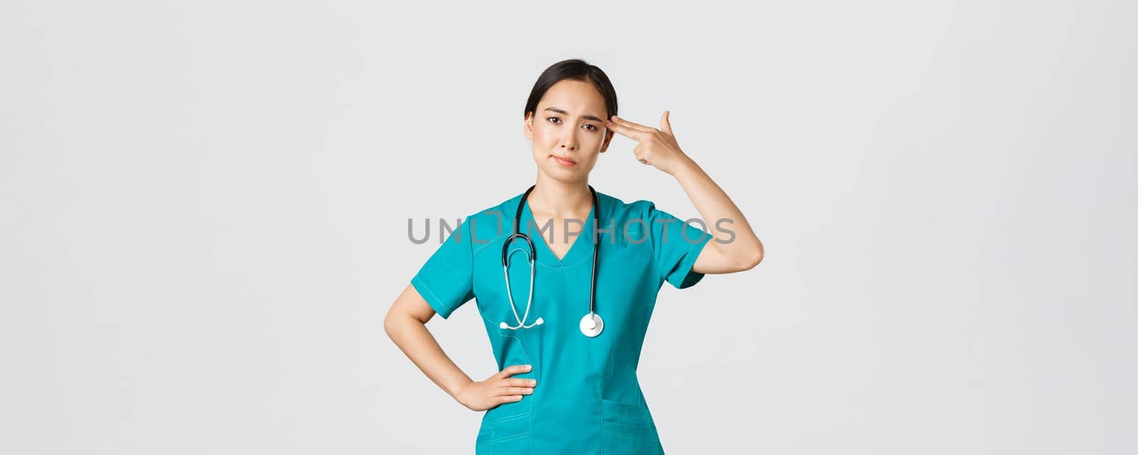 Covid-19, healthcare workers, pandemic concept. Exhausted and bothered, annoyed asian female doctor, nurse making shot gun gesture over head from boredom, white background.