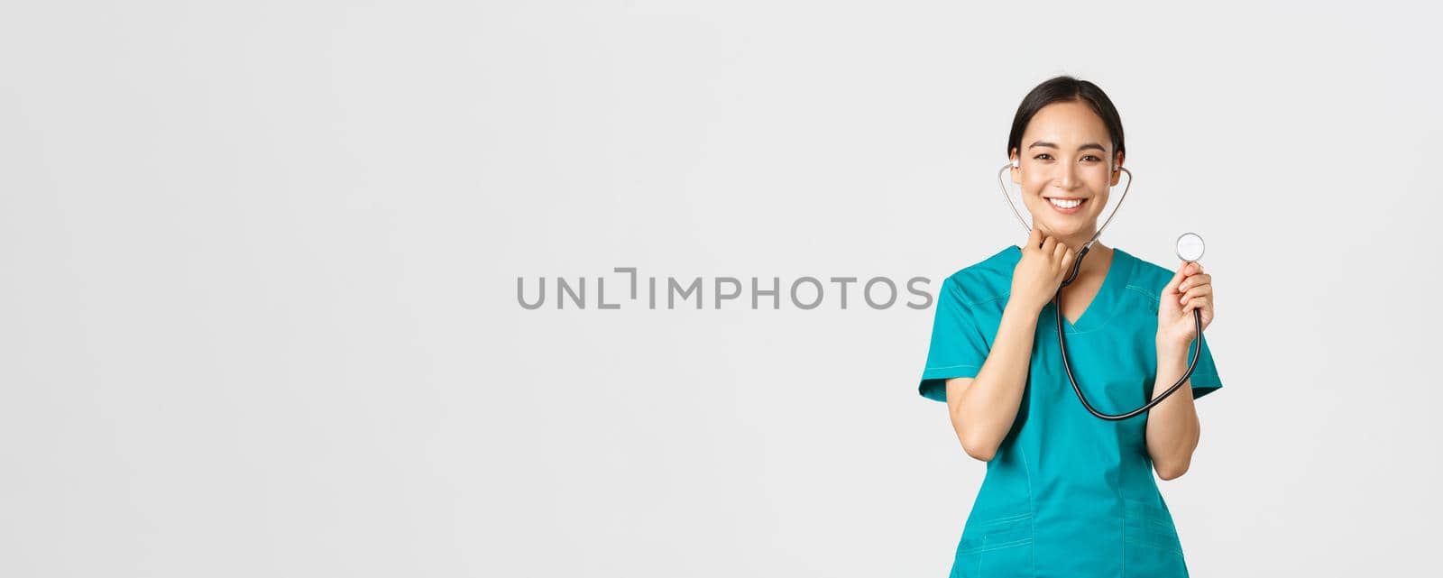 Covid-19, healthcare workers and preventing virus concept. Cheerful smiling asian female physician, doctor consult patient, examine person with stethoscope, standing white background.