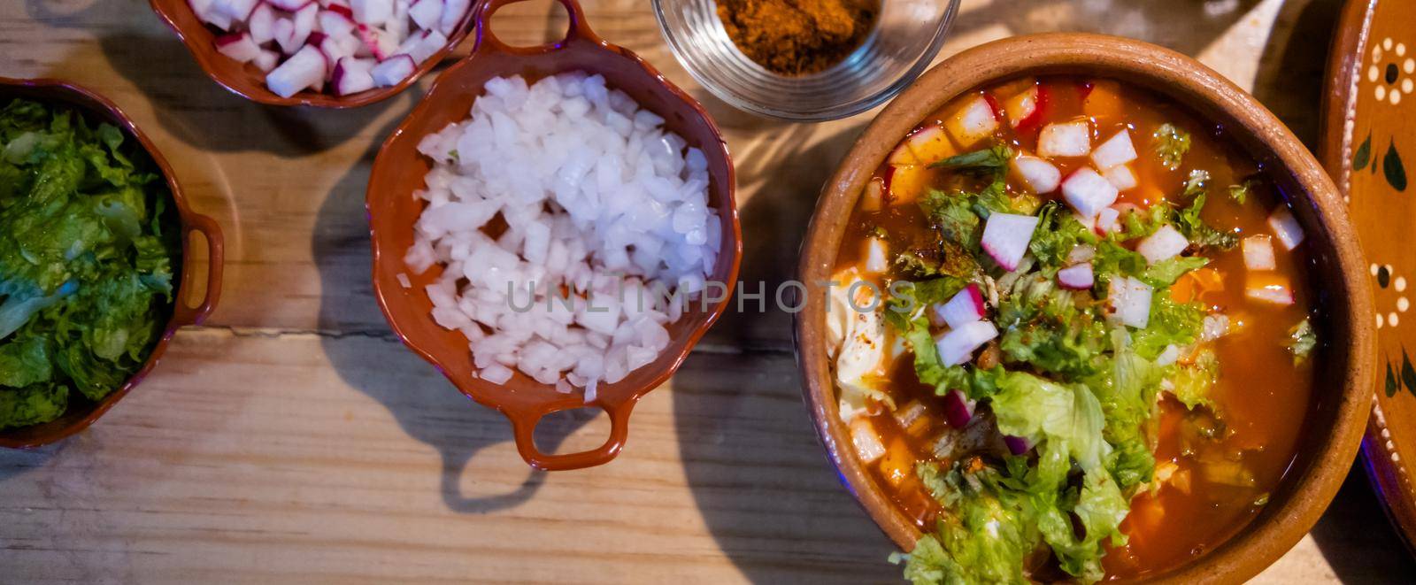 Wide view of bowl of delicious and traditional red pozole, chopped onions, sour cream, and tostadas from above. Authentic Hispanic pork stew and condiments on wooden table. Traditional Mexican cuisine