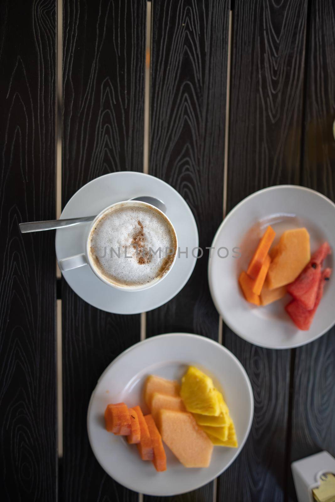 Top view of delicious cup of coffee latte, sliced fruit for breakfast on black table. Tasty hot drink and pineapple, melon, and papaya slices on white crockery above dark wooden surface. Balanced diet