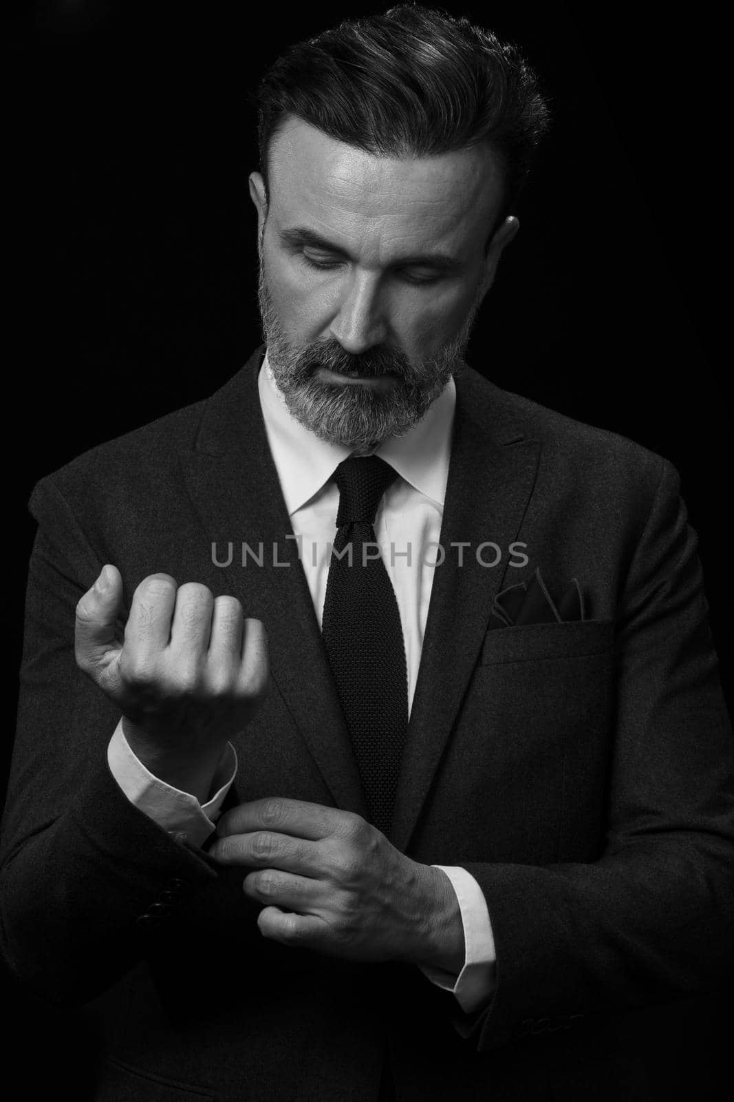 Portrait of a stylish elegant senior businessman with a beard and casual business clothes in photo studio isolated on dark background adjusting the suit