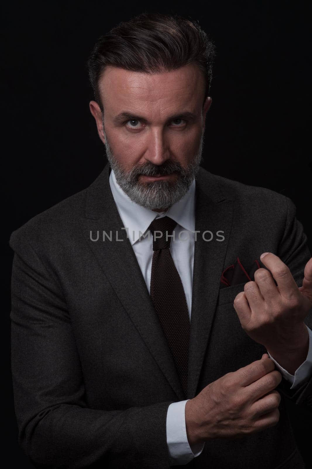 Portrait of a stylish elegant senior businessman with a beard and casual business clothes in photo studio isolated on dark background adjusting the suit