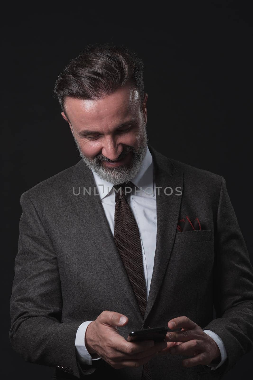Chat with client. Good looking young man in full suit using his smart phone and smiling while standing against grey background. by dotshock