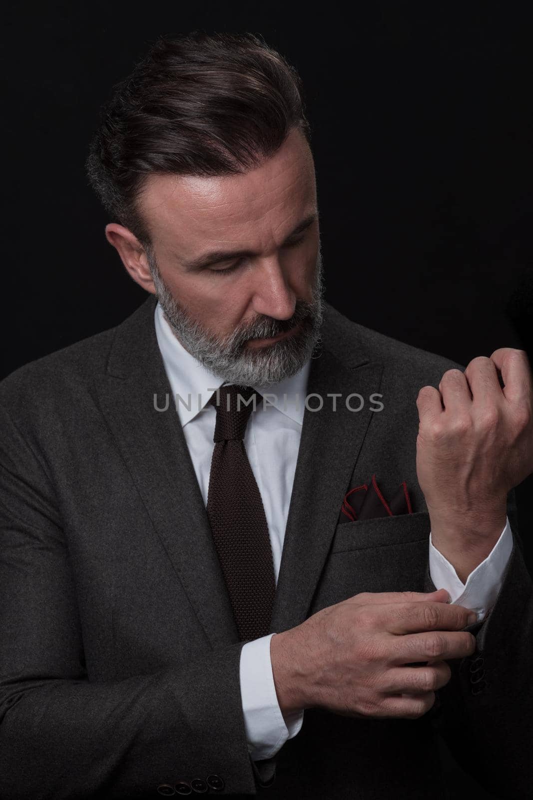 Portrait of a stylish elegant senior businessman with a beard and casual business clothes in photo studio isolated on dark background adjusting the suit