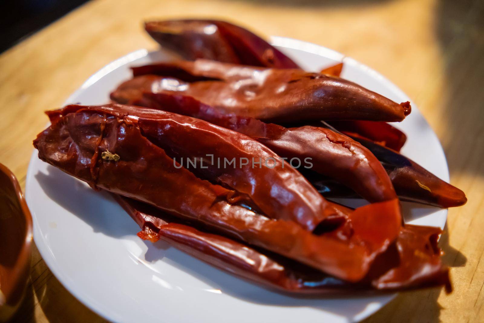 Close-up of group of dry red chili peppers on white plate above table. Several thin peppers on porcelain plate above wooden surface. Traditional hot sauce preparation