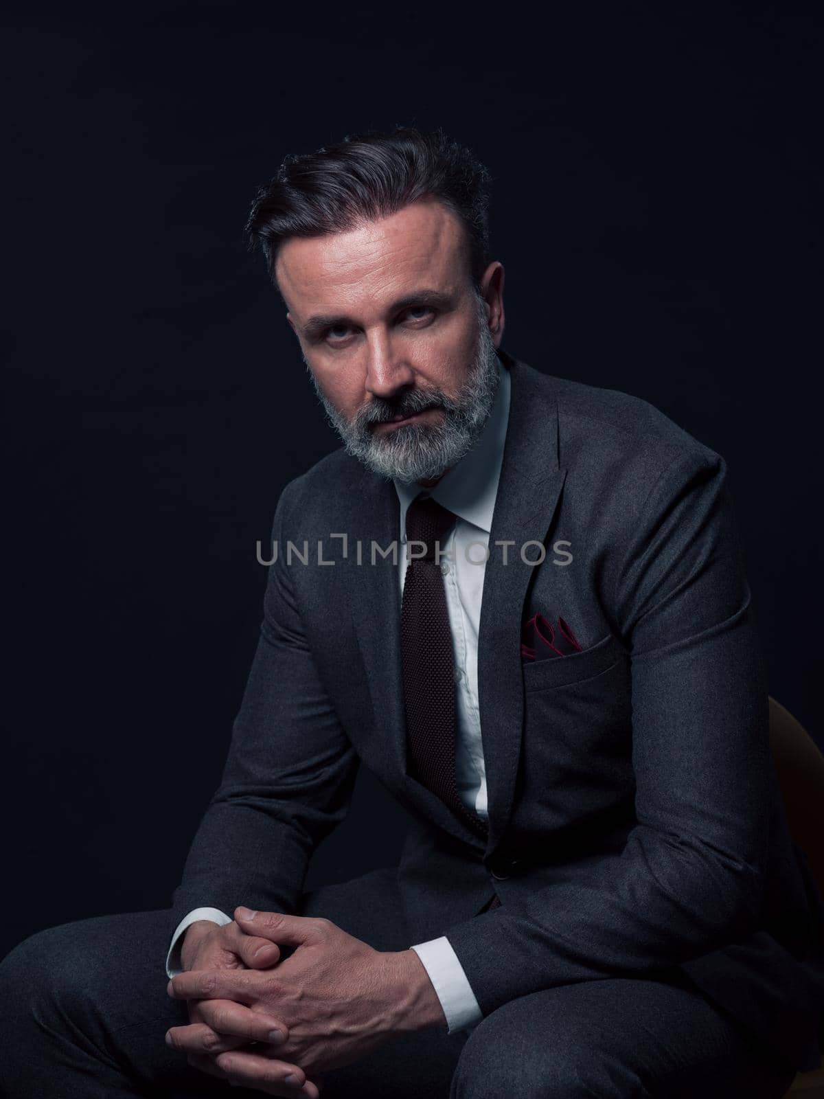 Portrait of adult businessman wearing trendy suit and sitting in modern studio on stylish chair against the black background by dotshock