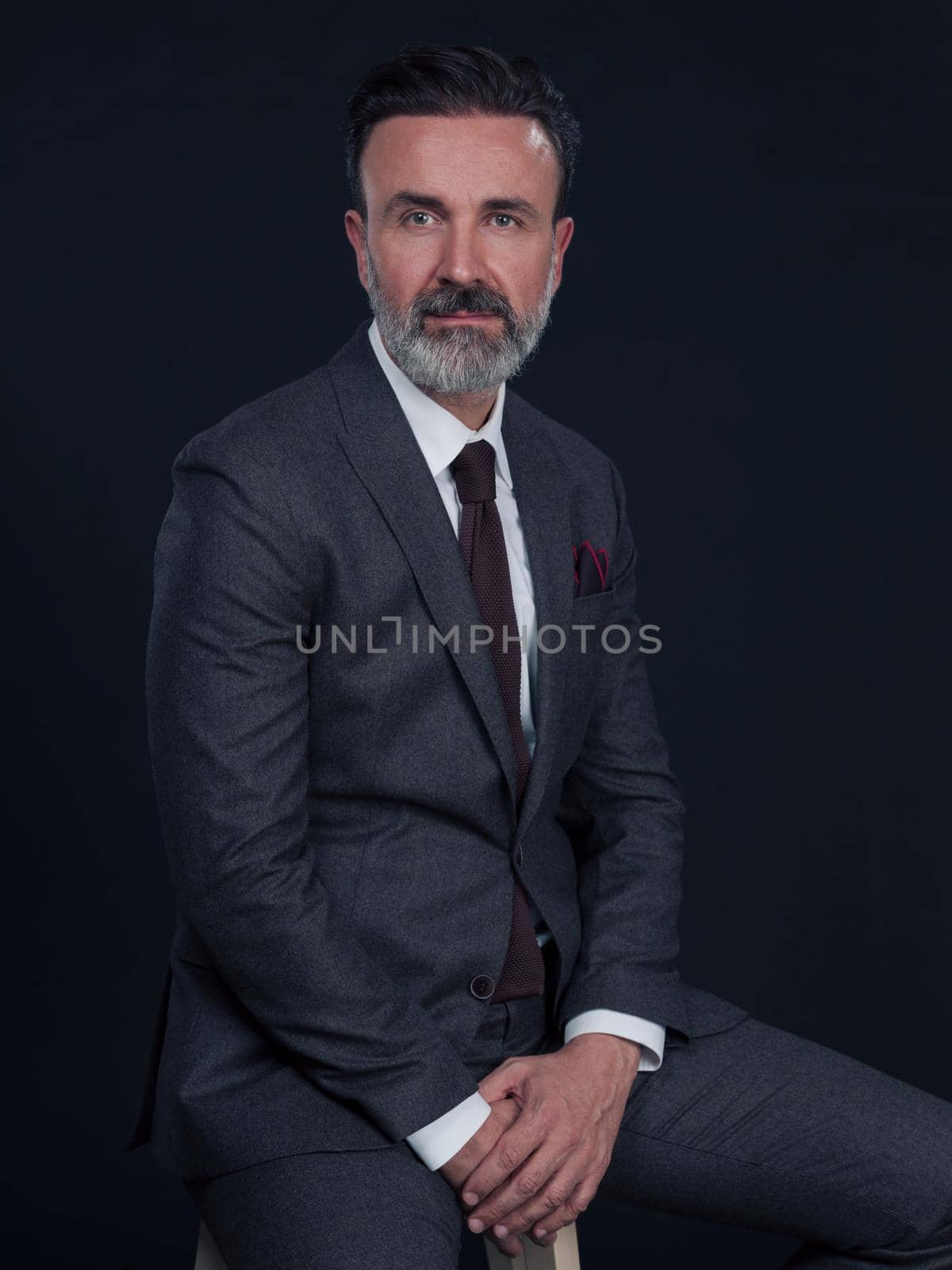 Portrait of adult businessman wearing trendy suit and sitting in modern studio on stylish chair against the black background. Horizontal mockup. High quality photo