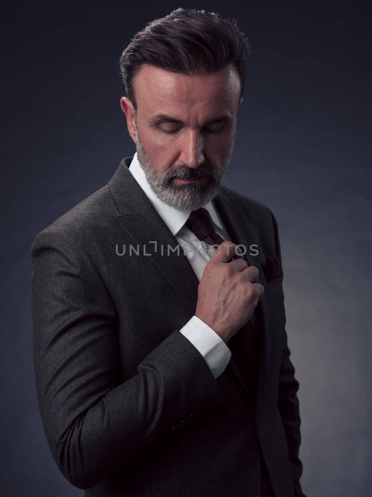 Portrait of a stylish elegant senior businessman with a beard and casual business clothes in photo studio isolated on dark background adjusting the suit