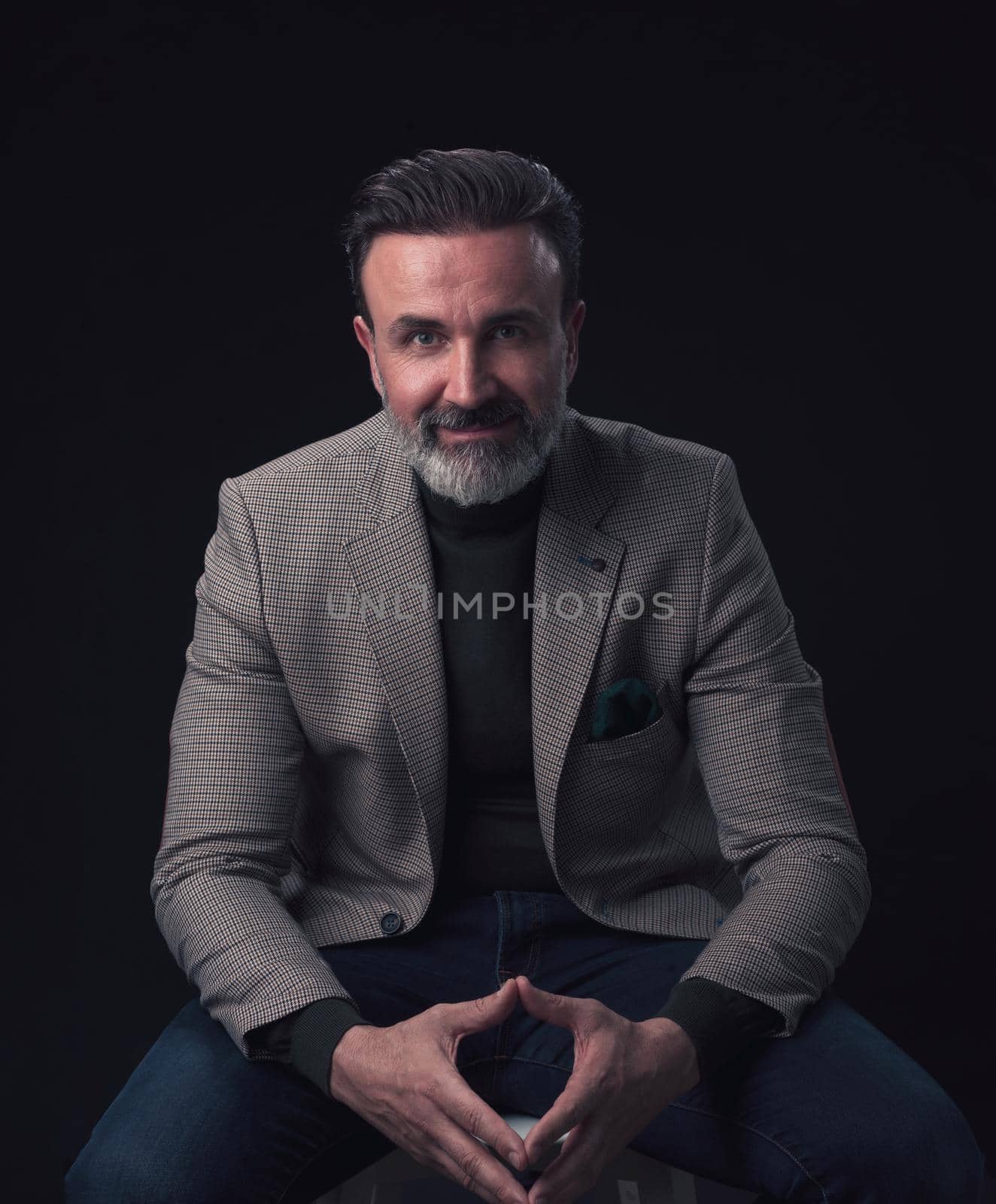 Portrait of adult businessman wearing trendy suit and sitting in modern studio on stylish chair against the black background. Horizontal mockup. High quality photo