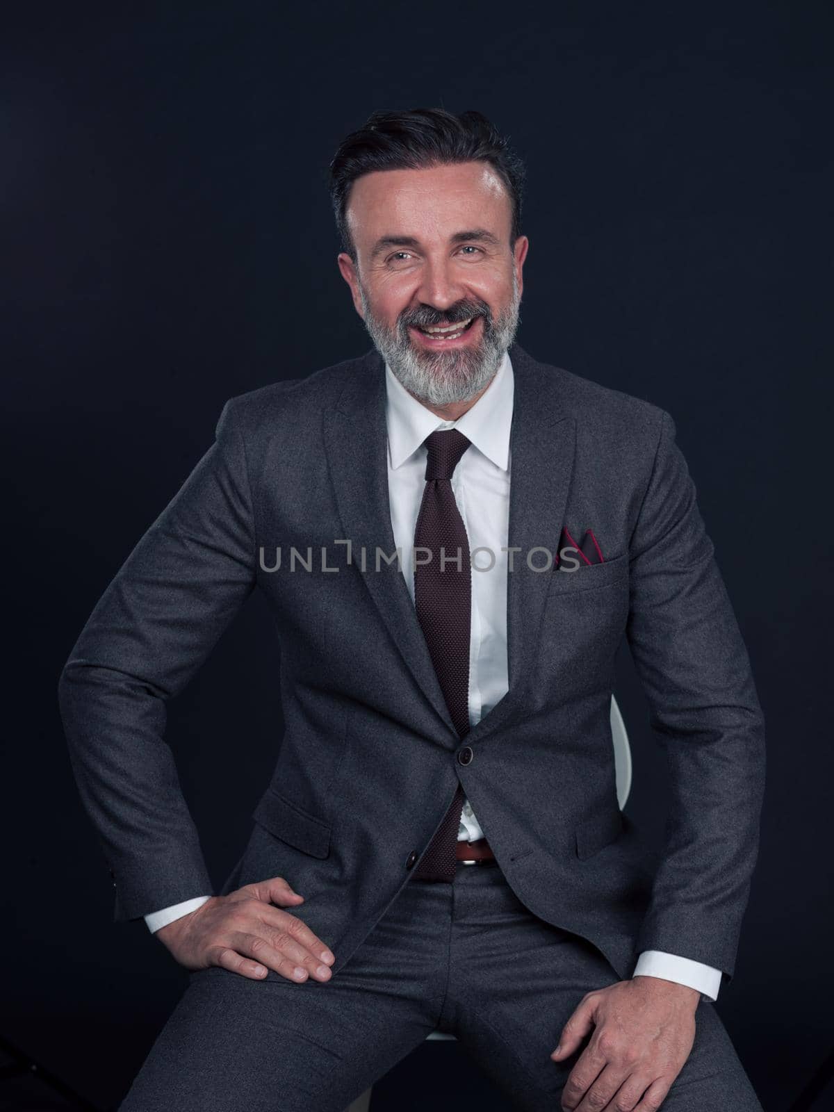 Portrait of adult businessman wearing trendy suit and sitting in modern studio on stylish chair against the black background by dotshock