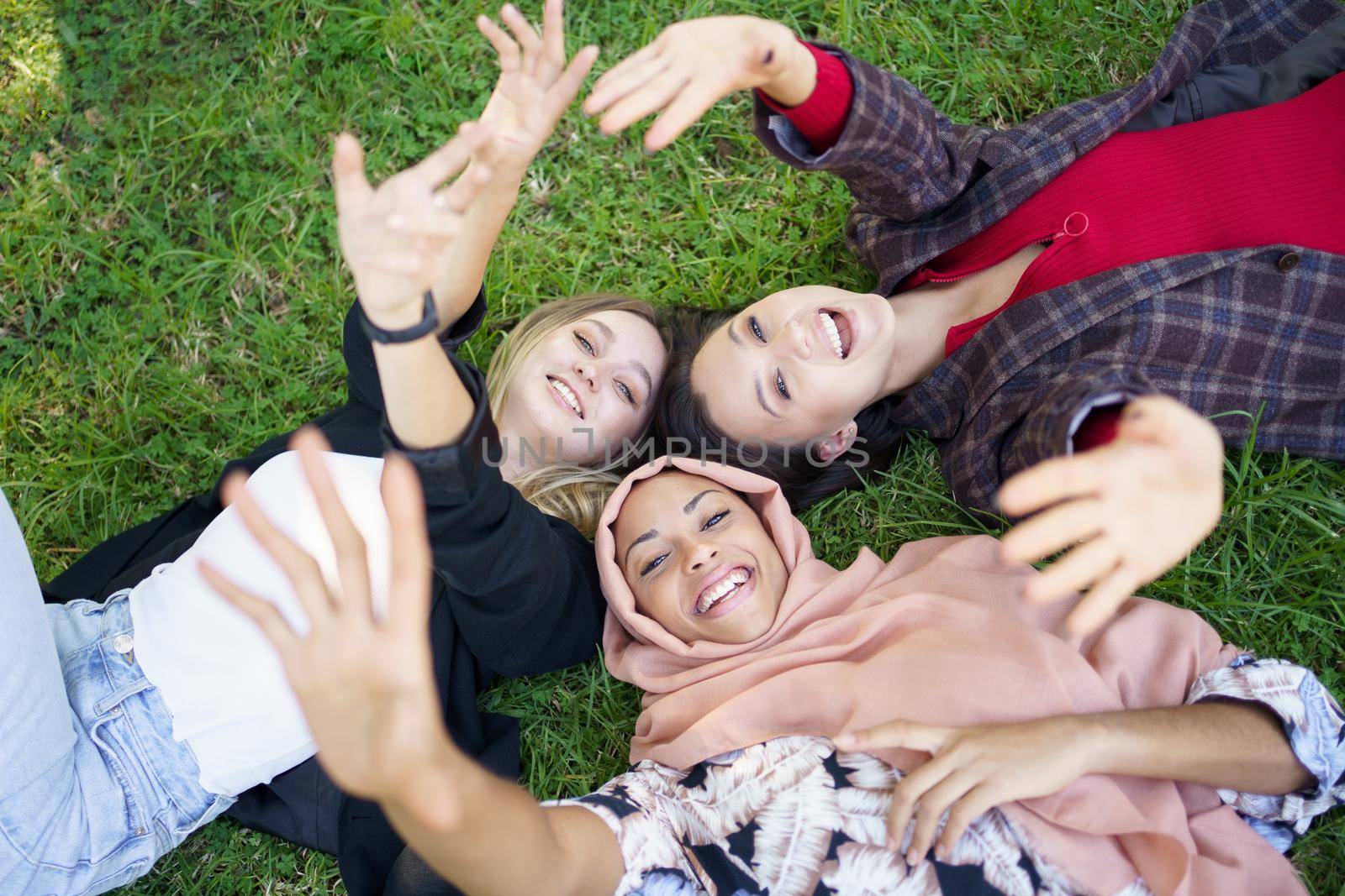 From above of happy young multiracial women smiling and waving hands at camera while lying on grassy meadow in park