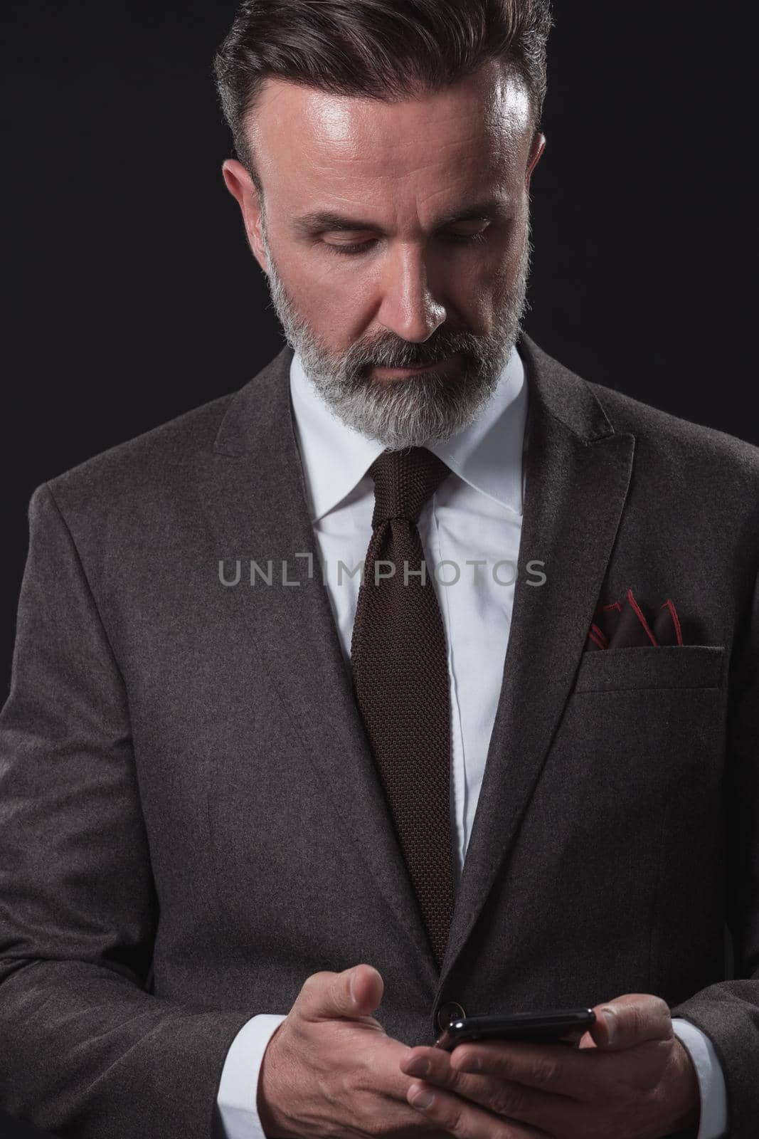 Chat with the client. Good looking young man in full suit using his smart phone and smiling while standing against dark grey background. High quality photo