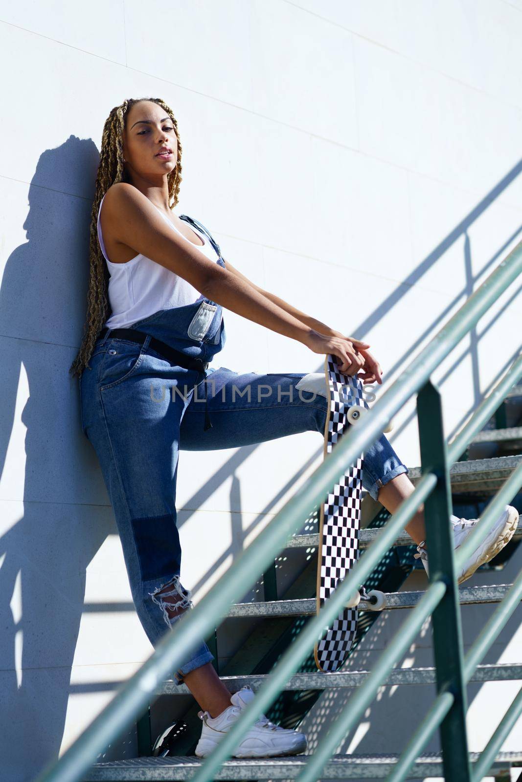 Young black girl with afro braids on an urban staircase, carrying a skateboard while looking at the horizon.