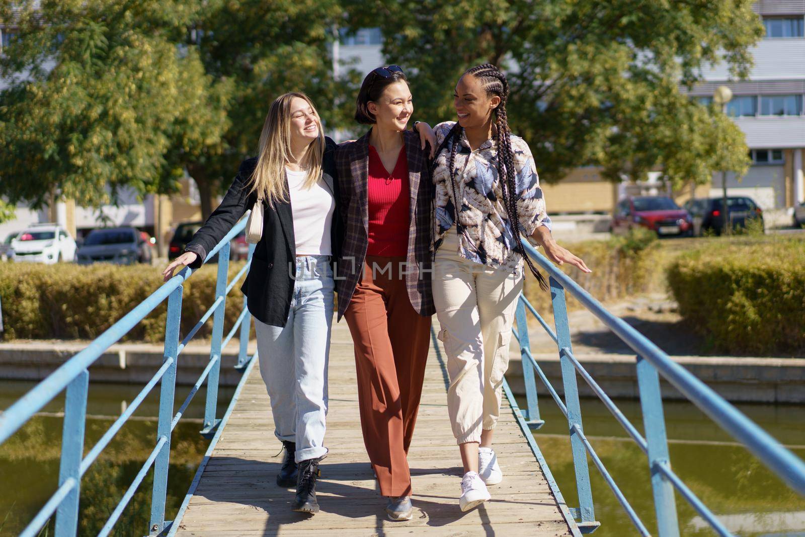 Smiling diverse friends hugging while strolling on narrow footbridge by javiindy