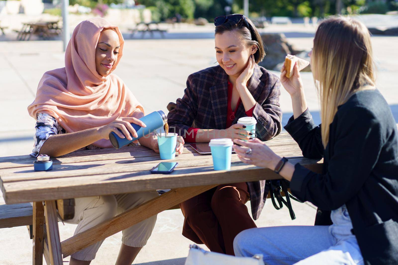 Happy young Muslim woman in hijab, pouring coffee from thermos into disposable cup, while having break with female friends in casual clothes sitting at wooden table in city park.