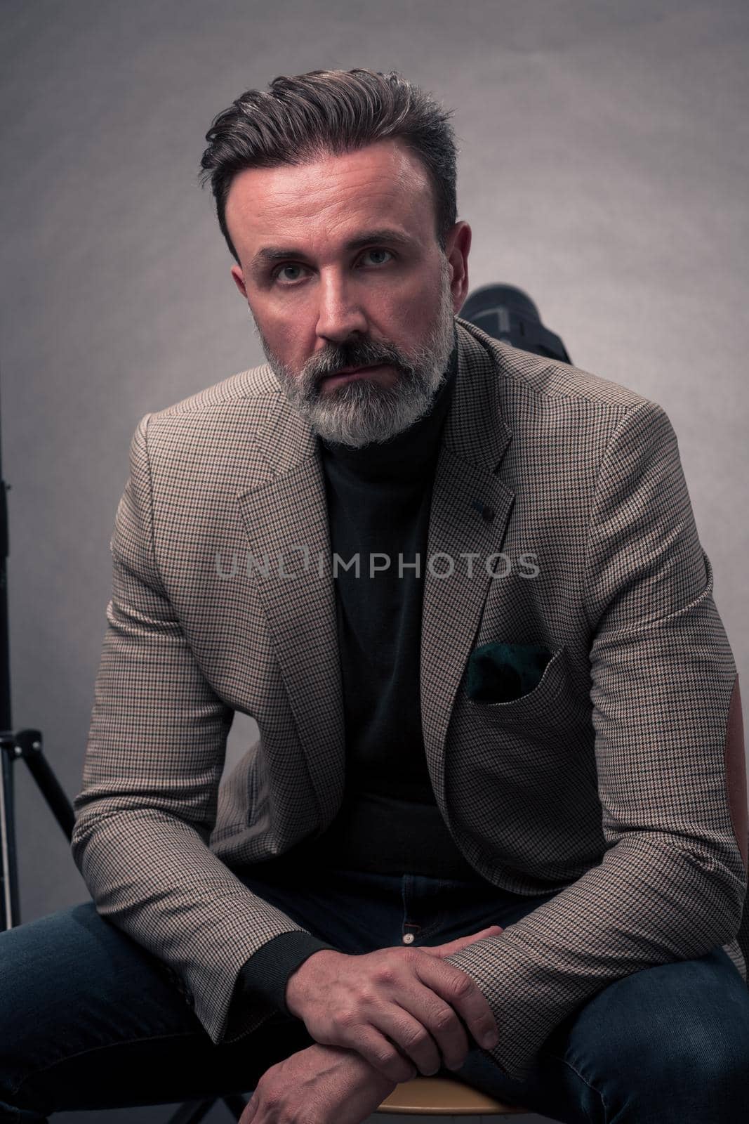 Portrait of adult businessman wearing trendy suit and sitting in modern studio on stylish chair against the black background. Horizontal mockup. High quality photo