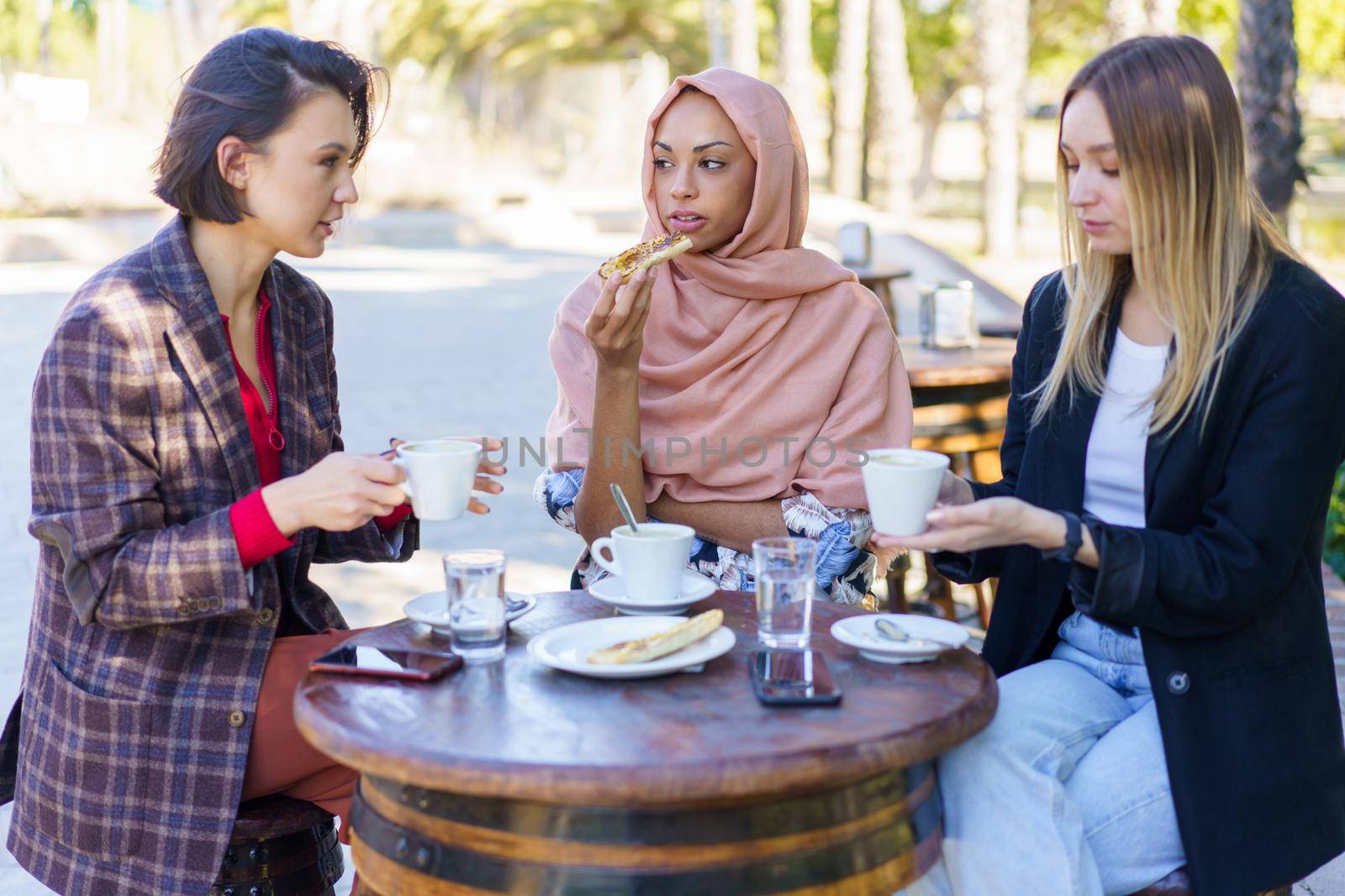 Stylish young multiracial female friends in casual clothes eating pastries and drinking coffee while spending time together in outdoor cafe