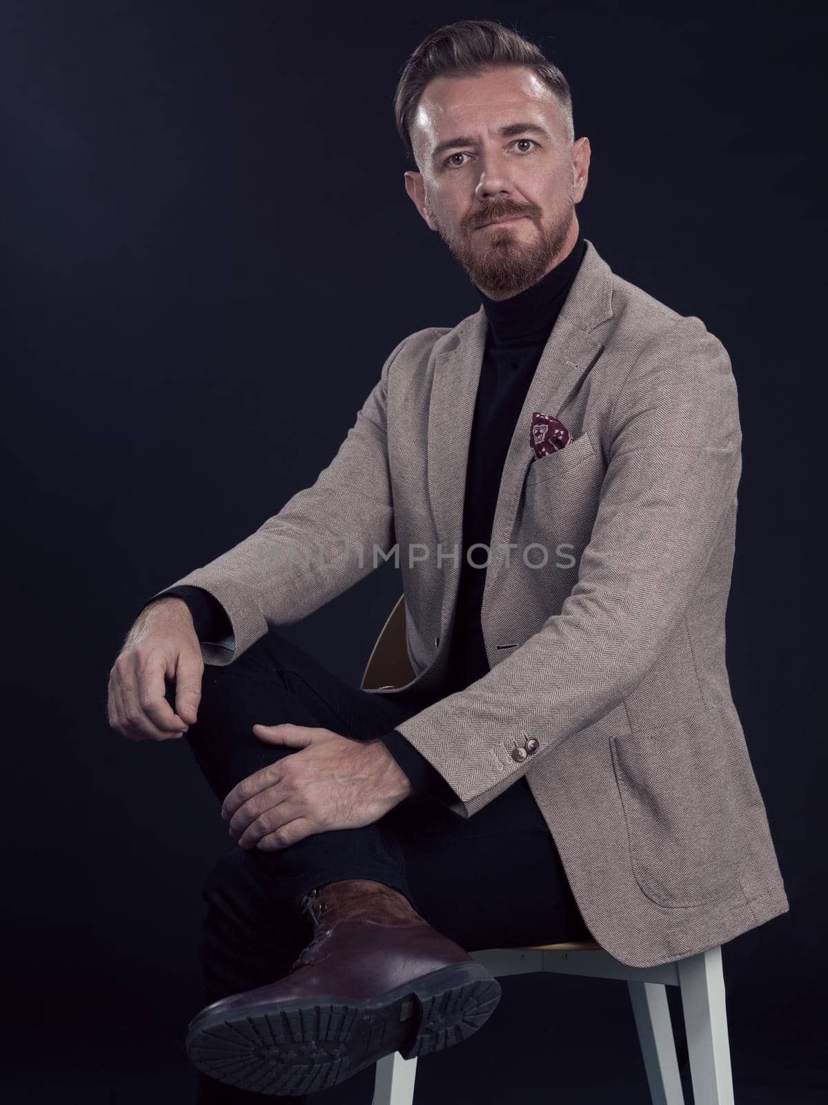 Portrait of adult businessman wearing trendy suit and sitting in modern studio on stylish chair against the black background. Horizontal mockup. High quality photo