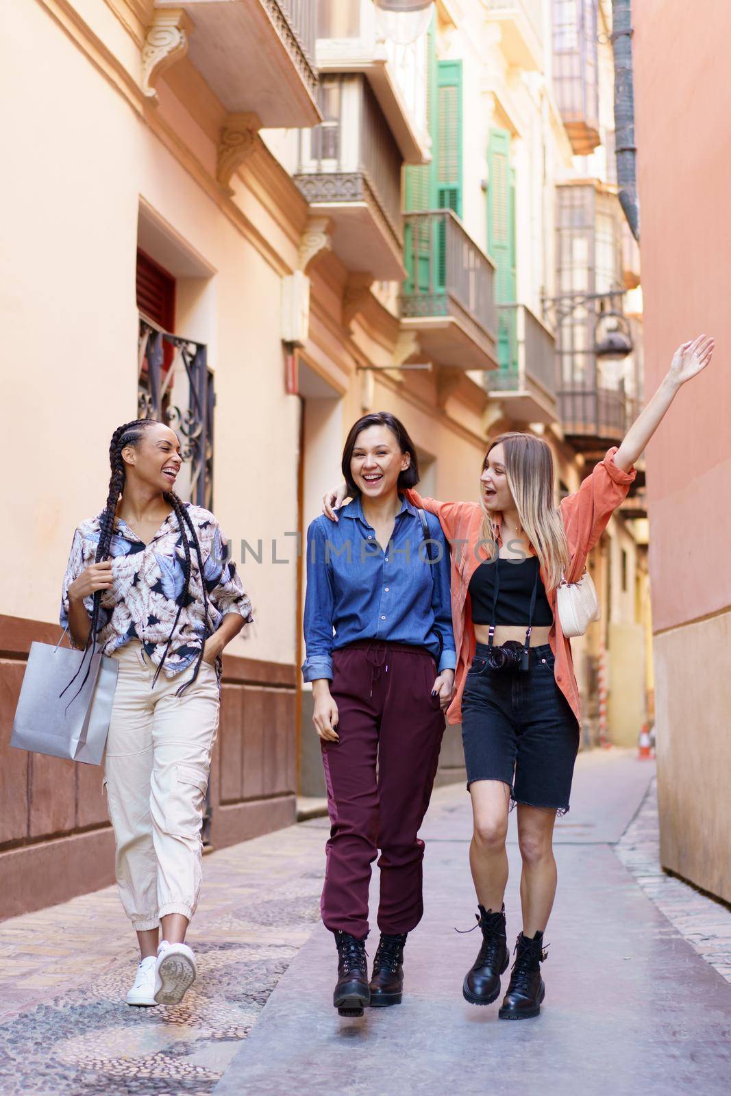 Full body of delighted young multiracial female, best friends in stylish clothes smiling and having fun together, while walking on narrow street between old buildings in city