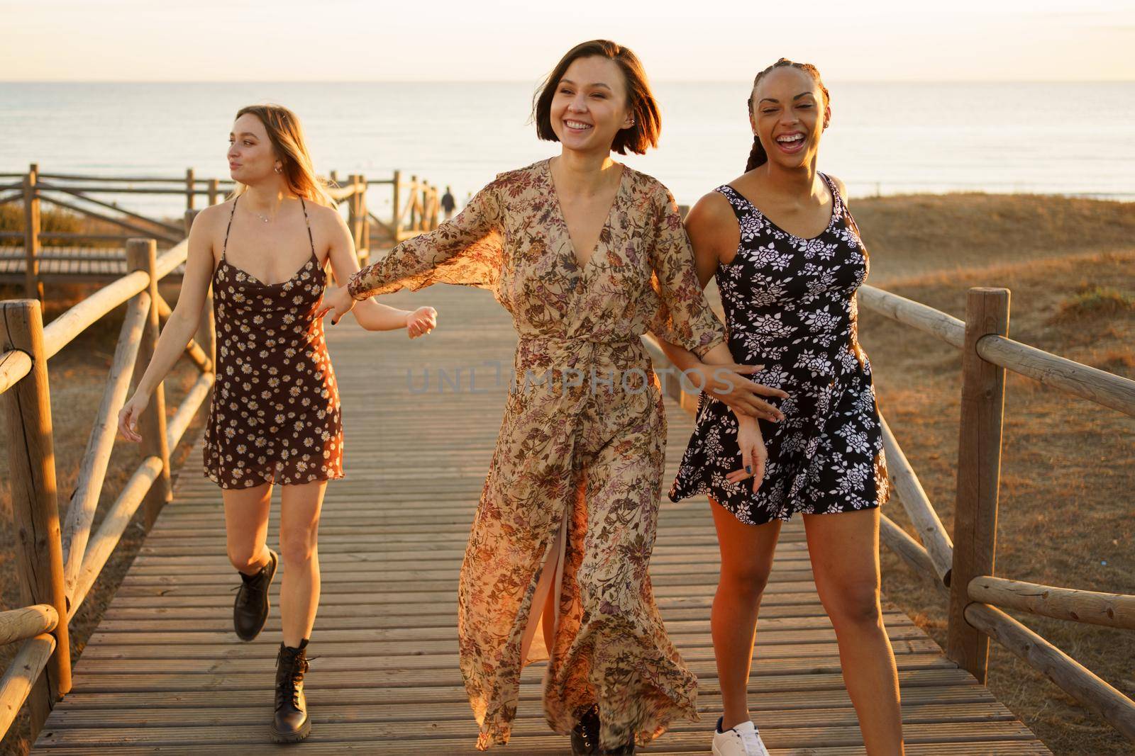 Smiling multiethnic girlfriends walking on wooden boardwalk on sea shore by javiindy
