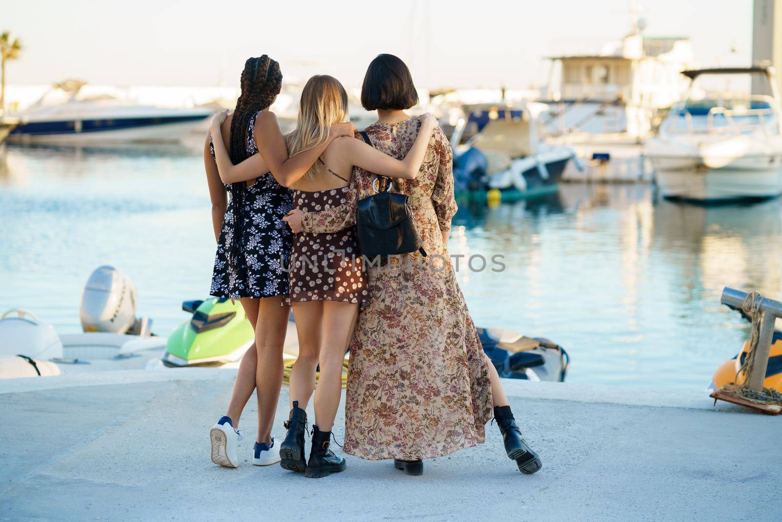 Full body back view of multiethnic unrecognizable female friends embracing gently while standing on seafront and observing marina