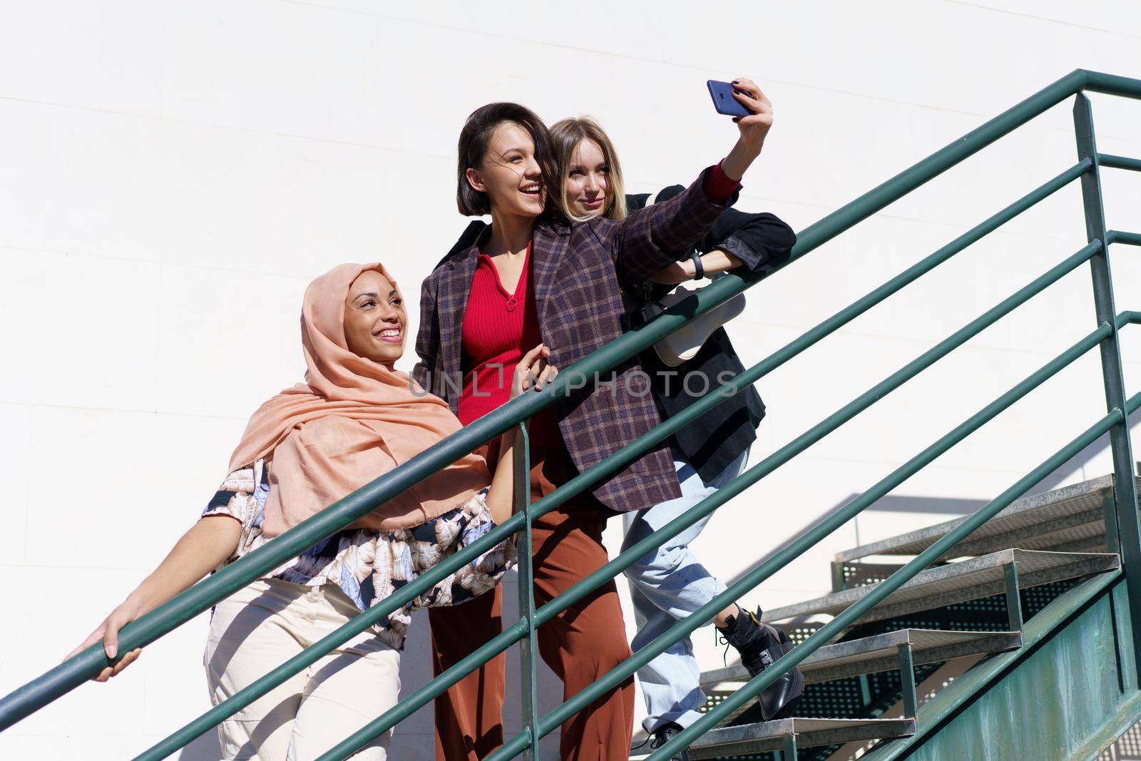 From below of positive young diverse female best friends smiling happily while taking selfie on smartphone standing on metal staircase in city on sunny day