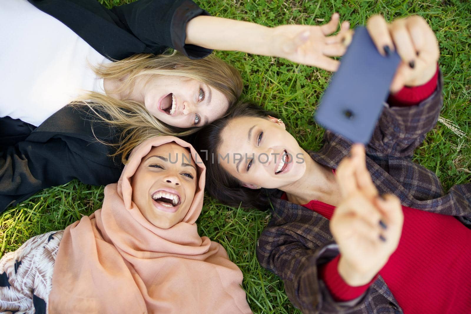 Cheerful diverse girlfriends laughing and taking selfie lying on lawn in park by javiindy