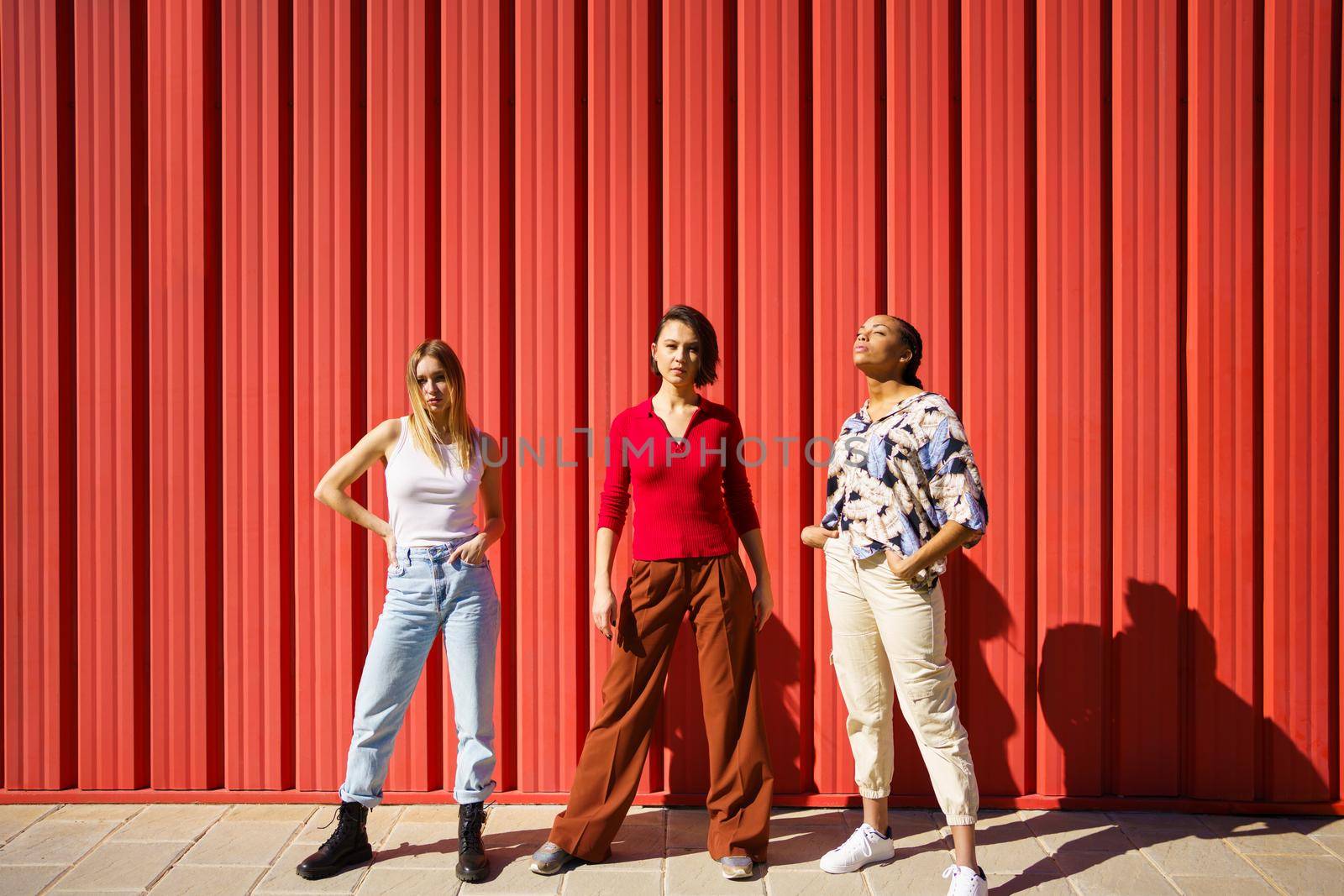 Full body of confident young multiracial female models in casual clothes standing near red metallic wall on sunny day in city