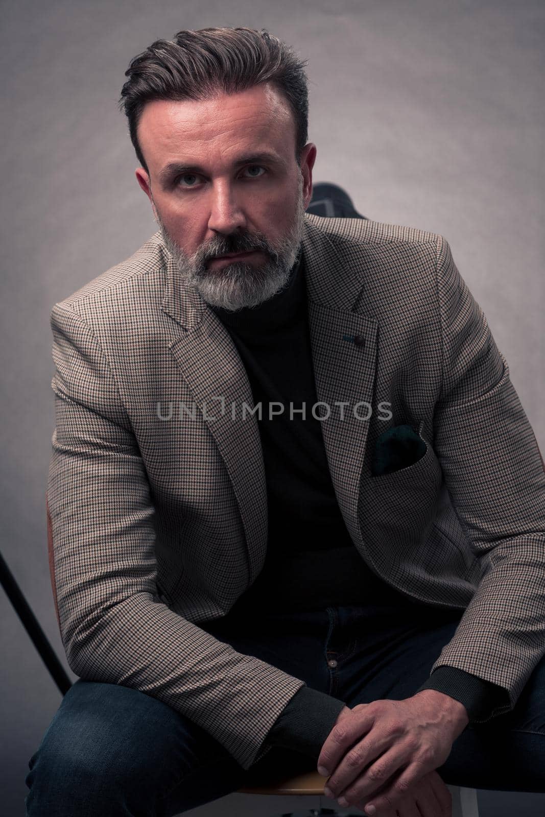 Portrait of adult businessman wearing trendy suit and sitting in modern studio on stylish chair against the black background. Horizontal mockup. High quality photo