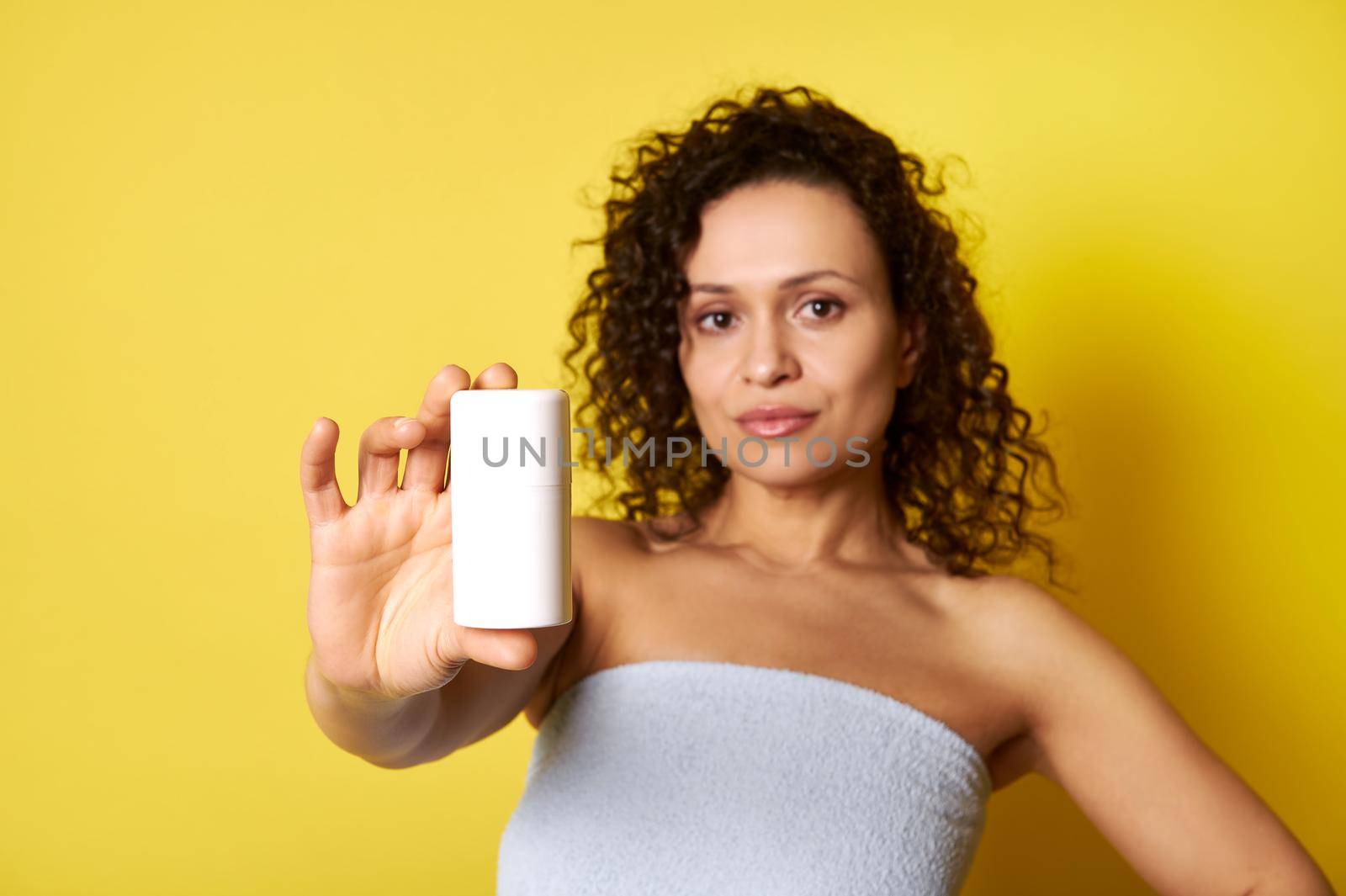 Soft focus on white container with cosmetic product in hand of beautiful half naked curly woman wrapped in bath towel. Body care concepts isolated on yellow background with copy space