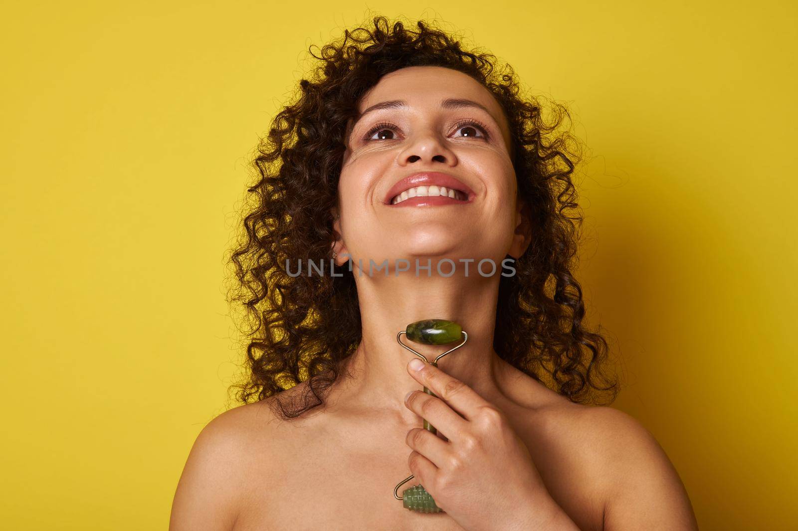 Smiling african american woman with curly hair massaging her neck with a jade roller, isolated over yellow background with copy space