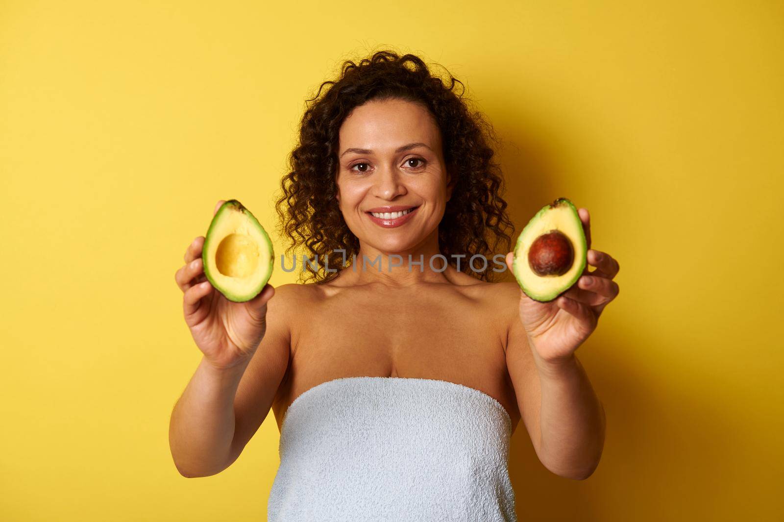 A curly-haired swarthy woman, wrapped in a bath towel holding avocado halves and showing them to camera, cute smiling with toothy smile while posing over yellow background with copy space