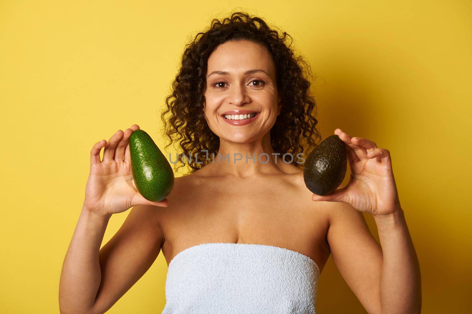 A curly-haired mixed race woman holding two ripe avocado fruits and smiling with toothy smile, isolated over yellow background with space for text