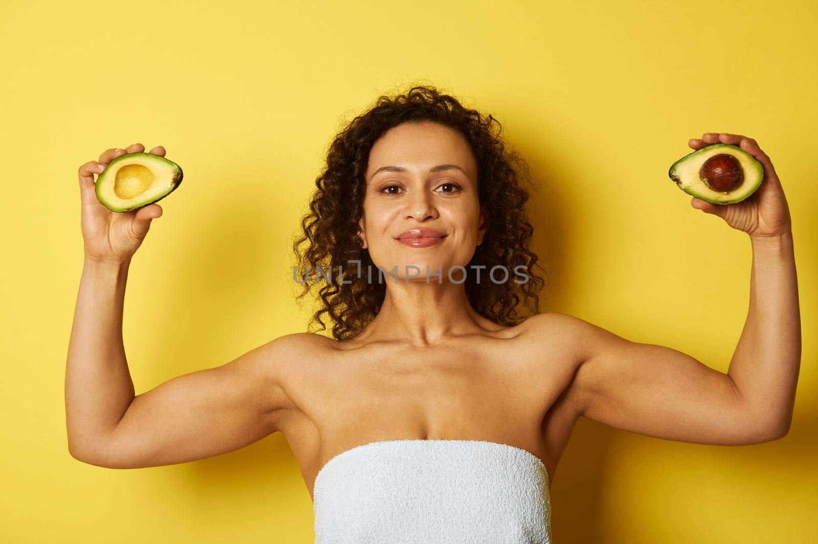 Young curly and muscular build African American woman wrapped in a bath towel holding two halves of ripe avocado, standing against yellow background with copy space