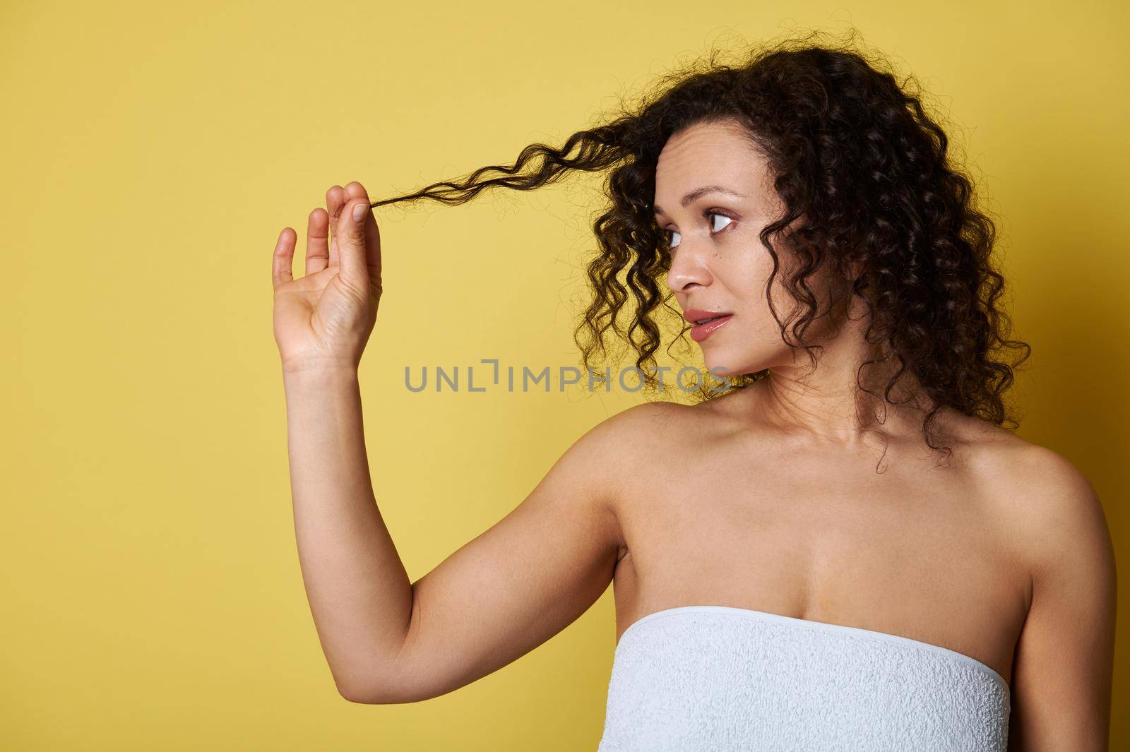 Young woman, wrapped in a bath towel, holding a lock of her curly hair and looking at her, posing against a yellow background with copy space