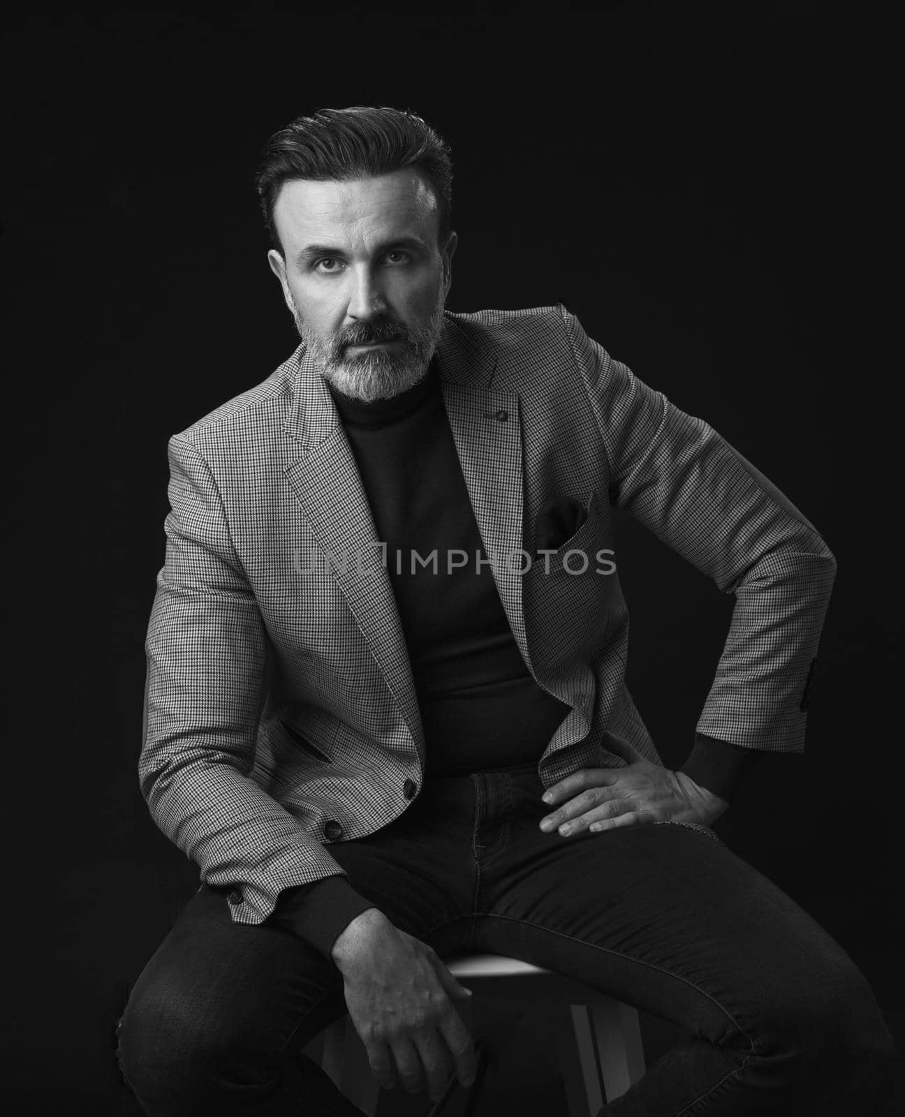 Portrait of adult businessman wearing trendy suit and sitting in modern studio on stylish chair against the black background. Horizontal mockup. High quality photo