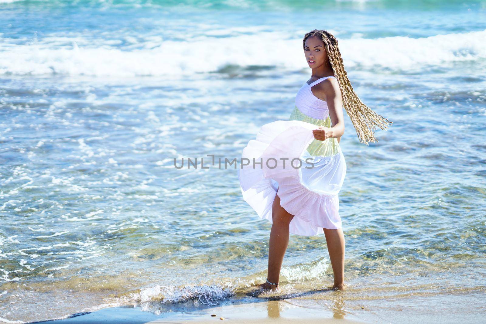 Black woman walking along the shore of the beach wearing a beautiful long dress. Young girl enjoying her vacations.