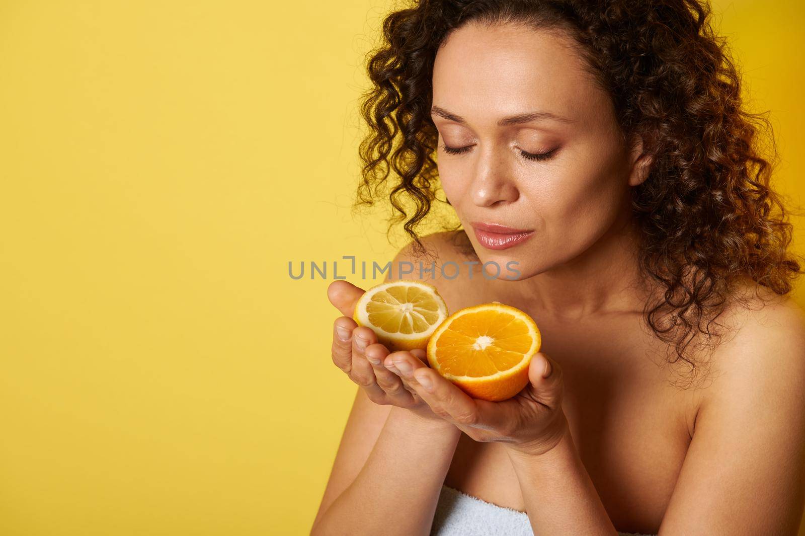Beauty portrait of attractive curly half-naked woman with natural makeup and glowing hydrated facial skin, enjoying citrus scent in her hands. Isolated over yellow background with copy space