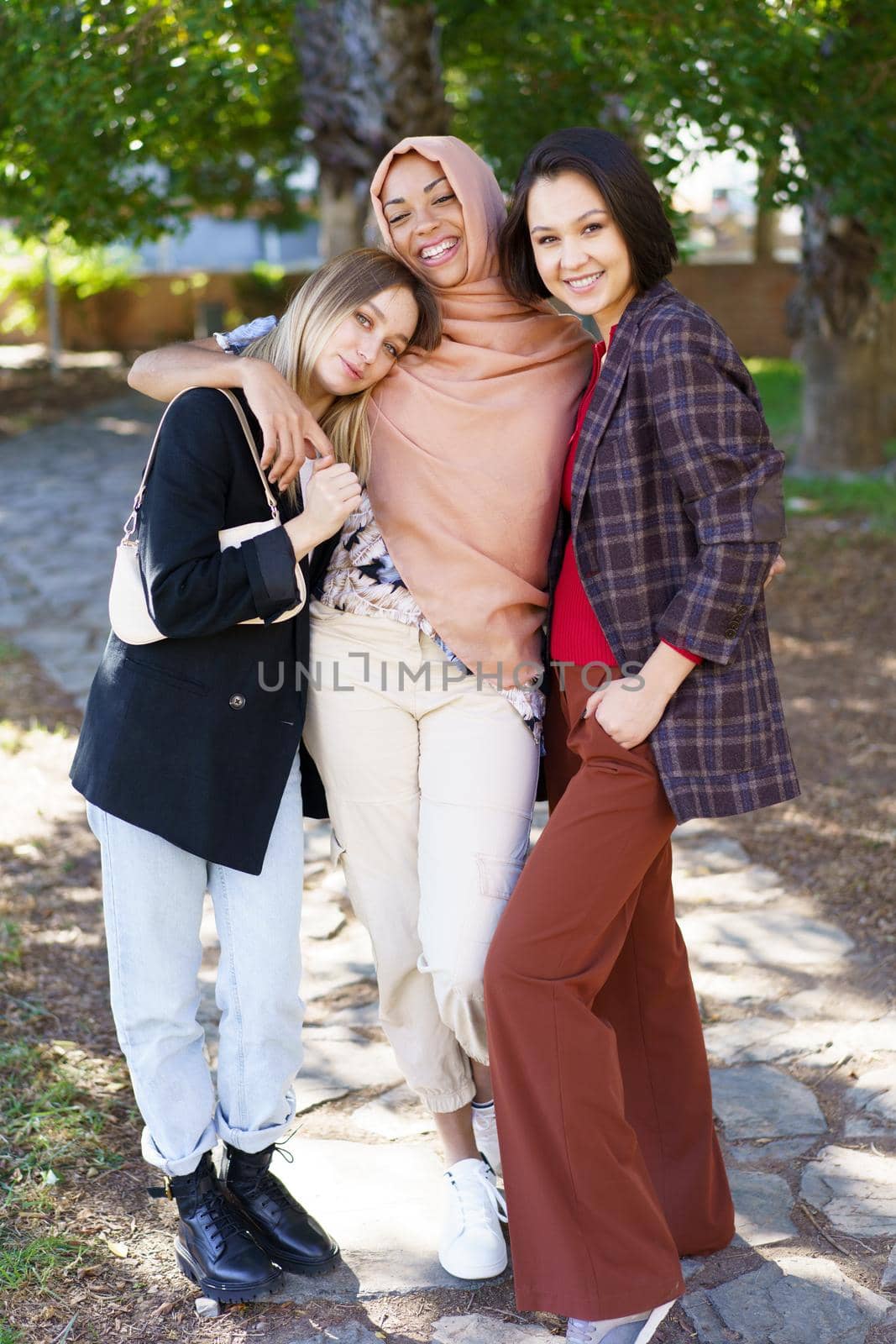 Positive young multiethnic female best friends in casual clothes smiling and looking at camera while cuddling in park on sunny day