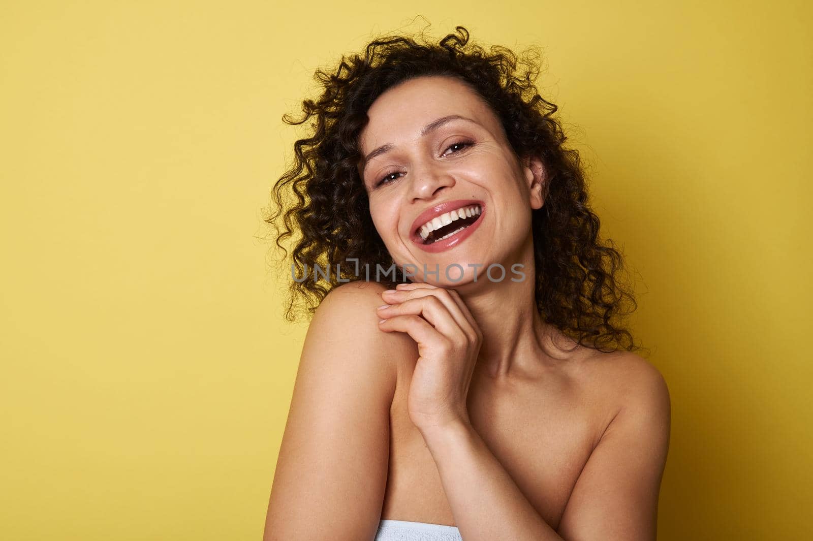 Beauty portrait of young half naked woman with curly hair smiling toothy smile looking at camera, posing over yellow background with copy space