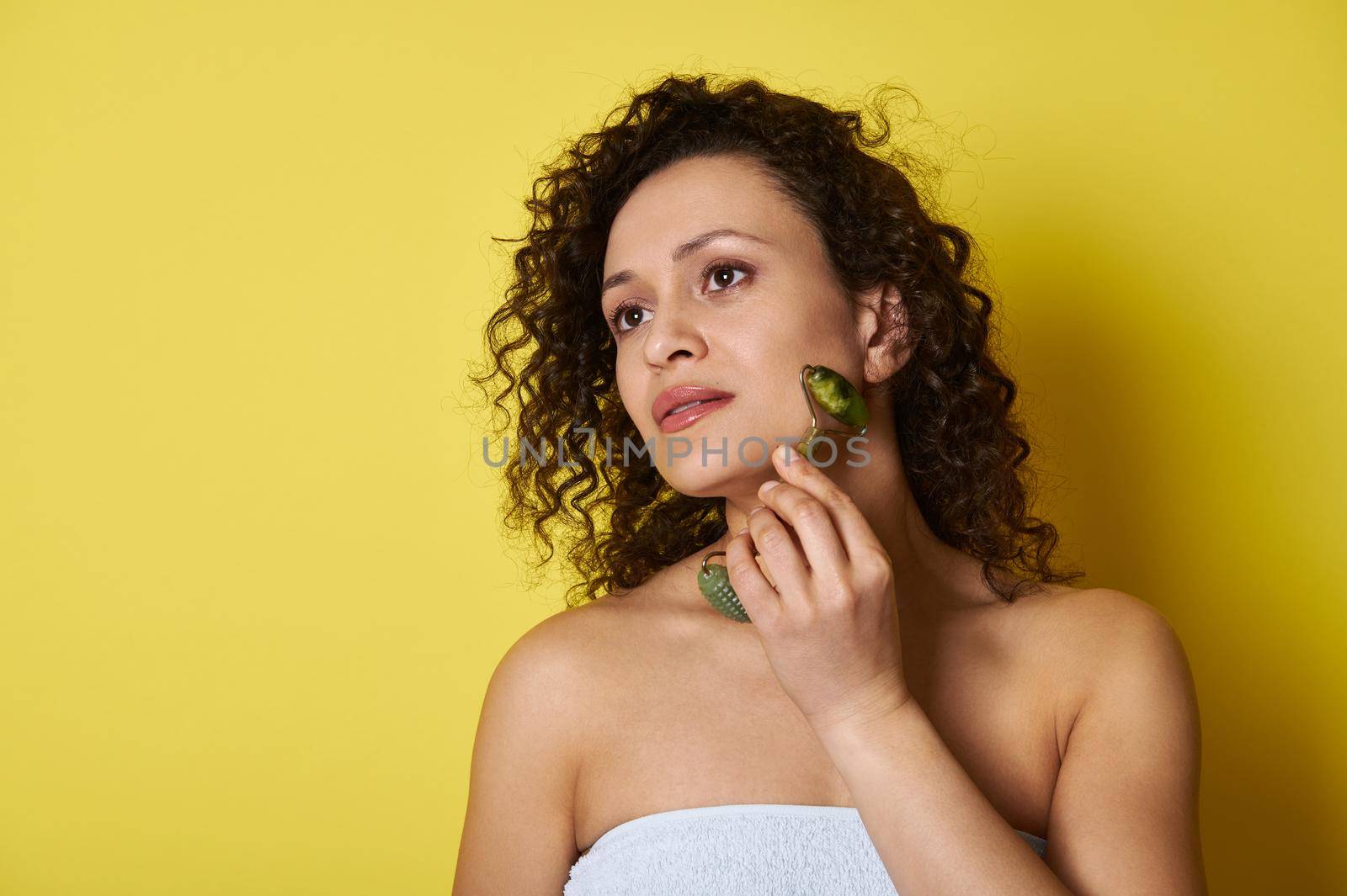 Beautiful young woman with curly hair using jade roller for facial massage and skin care. Shot with soft shadow over yellow background with copy space.