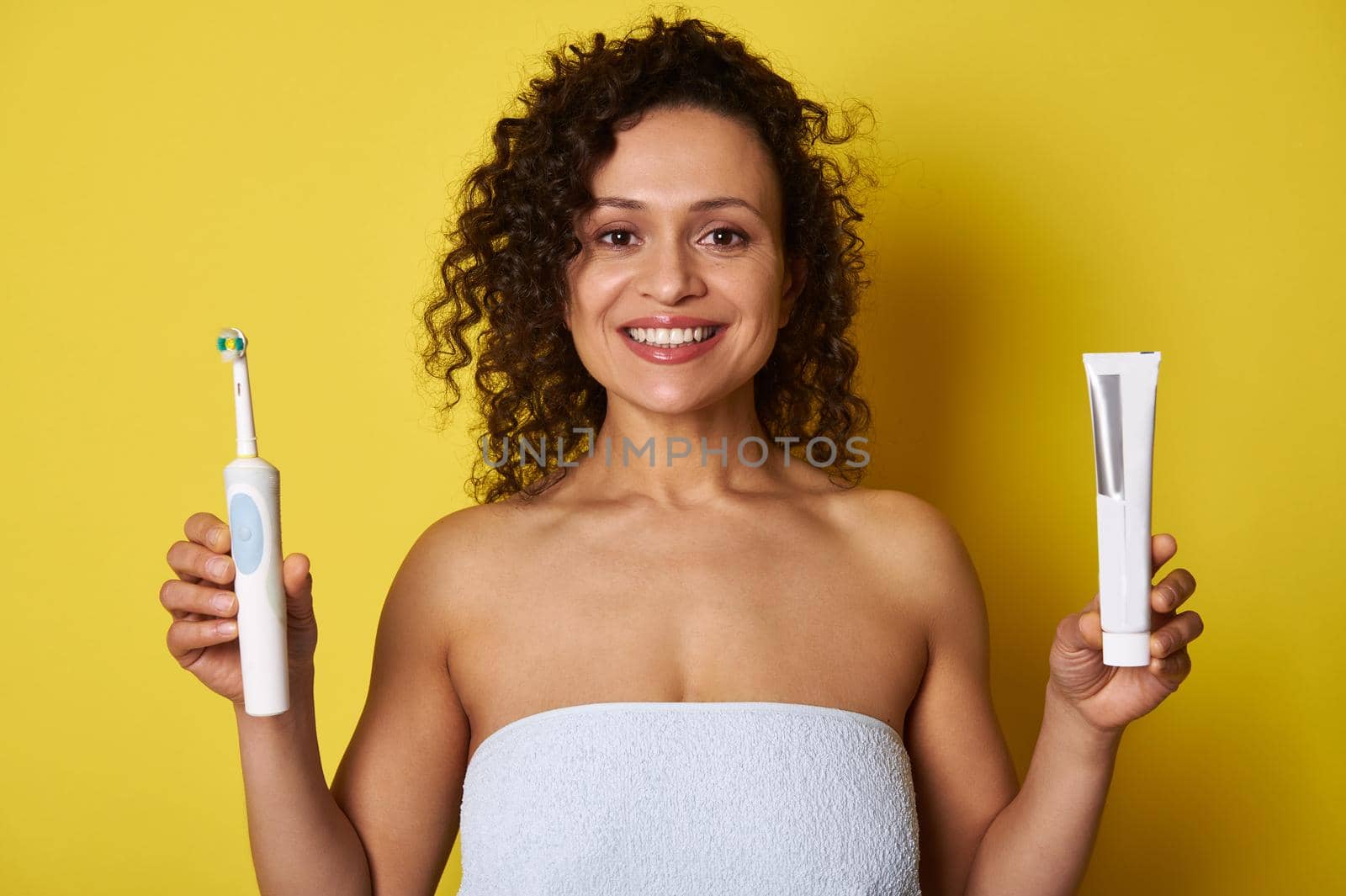 Beautiful half naked woman wrapped in a white bath towel holding a toothpaste in one hand and an electric toothbrush in the other hand and looking at the camera with toothy smile