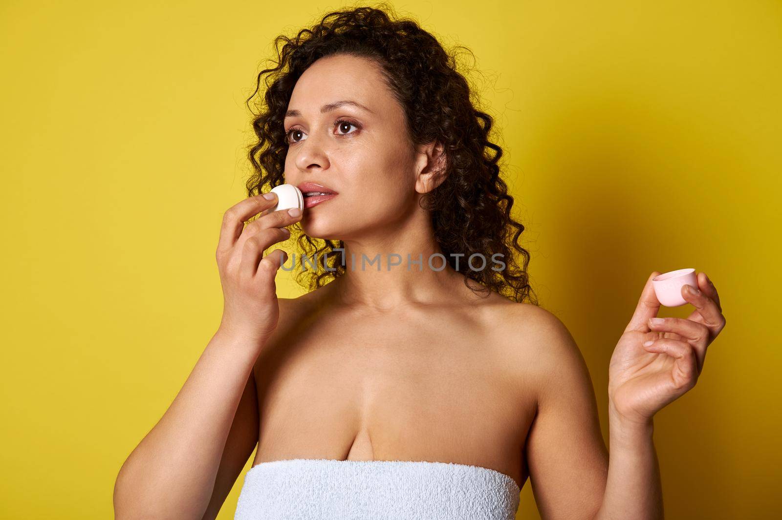 A young woman wrapped in a bath towel using a hygienic lipstick for hydrating and taking care about her lips. Copy space, yellow background