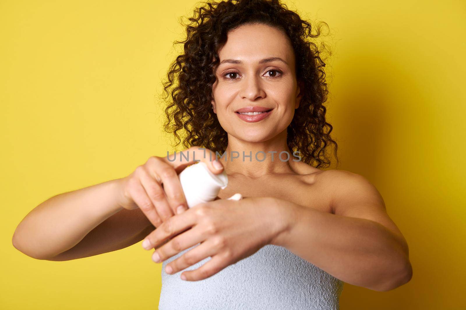A middle-aged curly woman squeezing moisturizing cream from a bottle and applying it on hands, isolated over yellow background with space for text