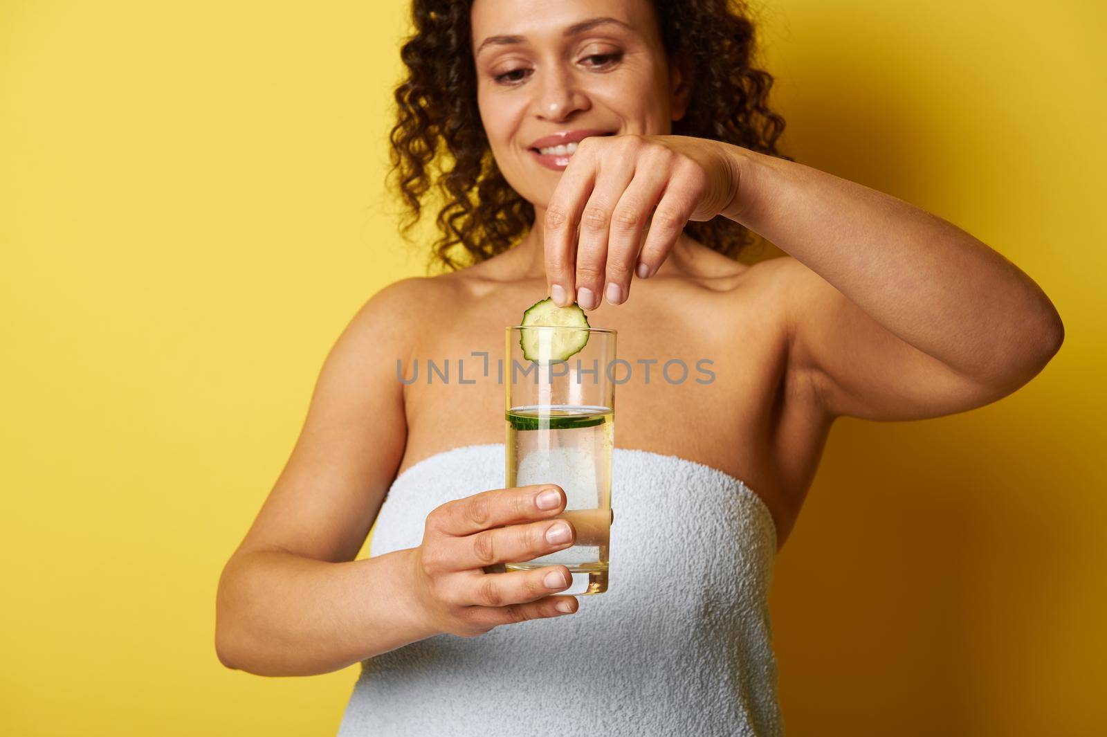 Closeup of young African woman's hand throwing a slice of cucumber into a glass with water by artgf