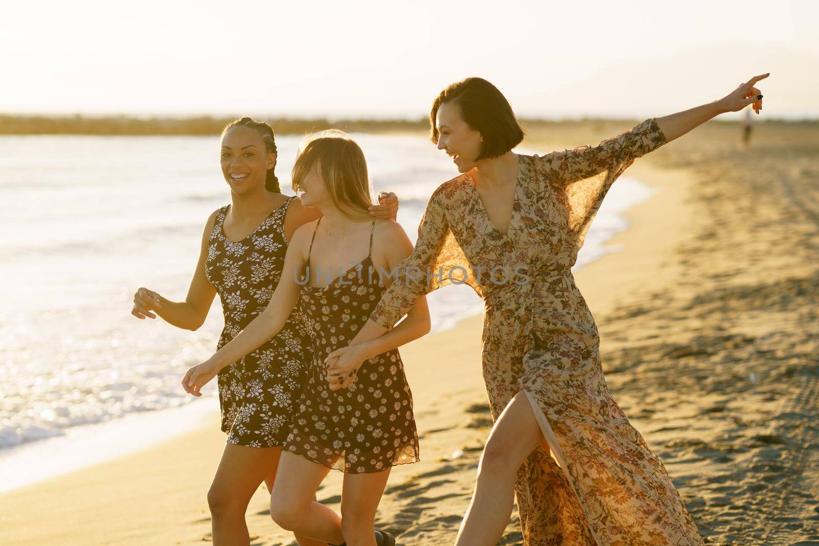 Happy multiethnic girlfriends in summer dresses holding hands and hugging while walking on sandy beach near sea in sunny evening