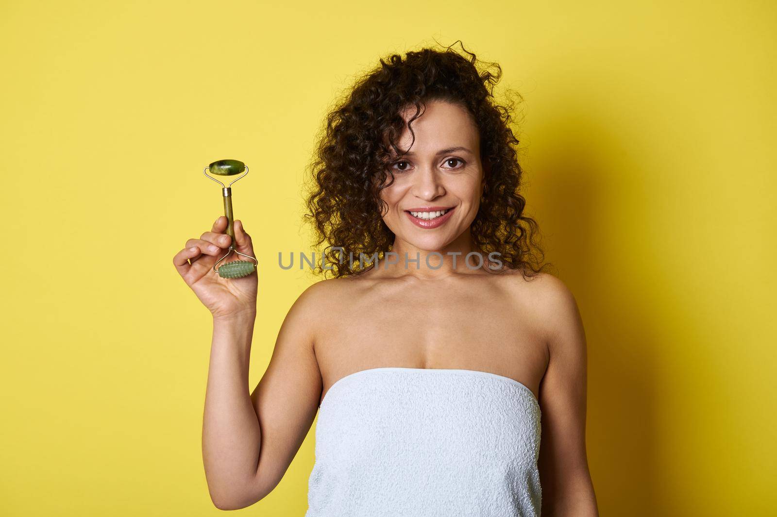 Beauty portrait of young curly haired woman holding jade roller and smiling toothy smile posing over yellow background with copy space