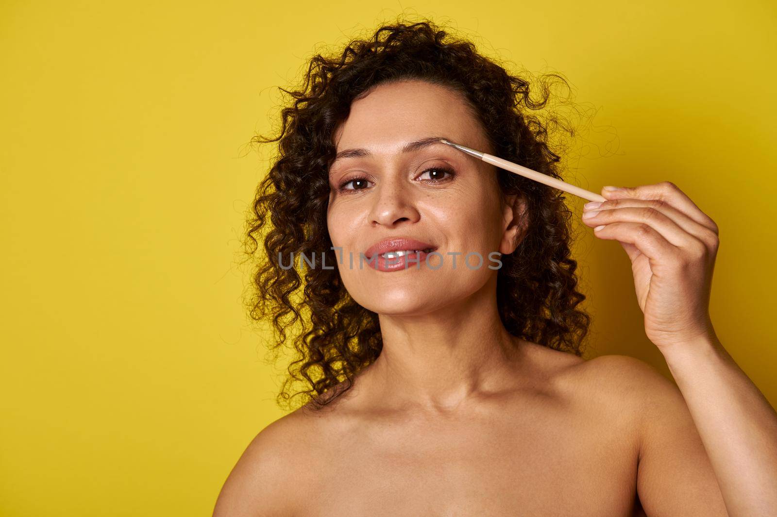 Smiling dark skin woman using makeup brush to correct and dye eyebrows. Make-up concepts on yellow background with copy space