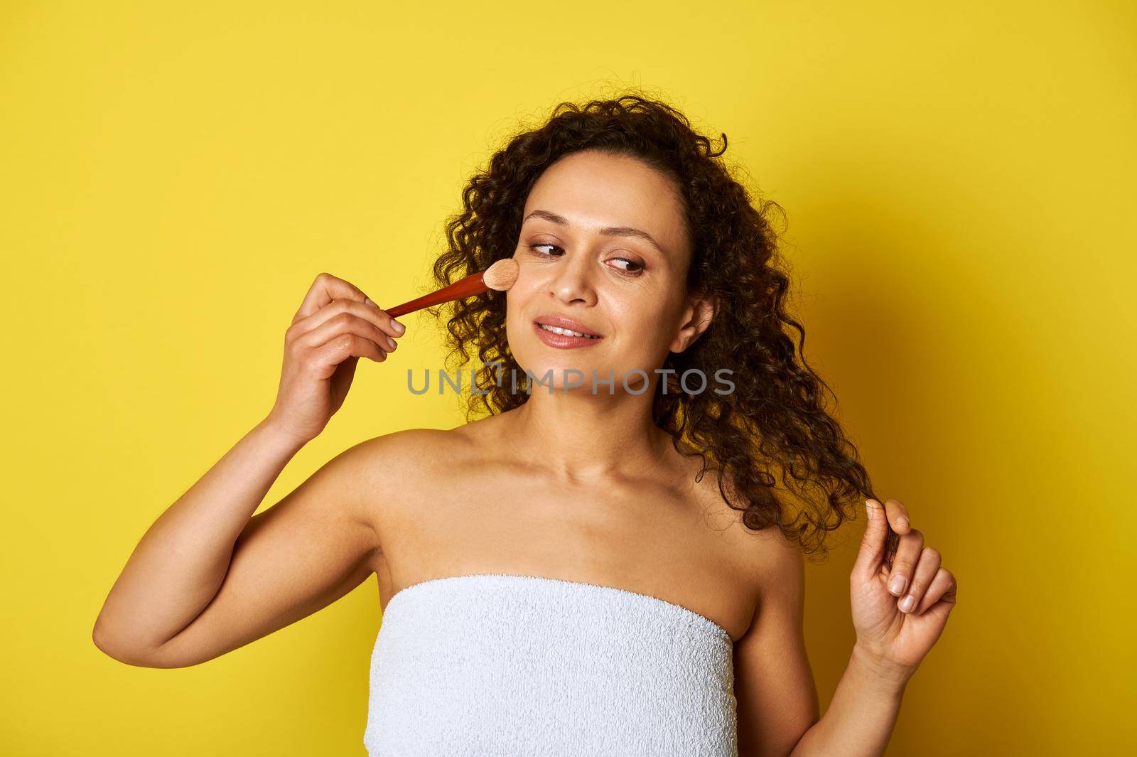 Charming half naked mulatto woman holding a makeup brush and applying blush on her face. Isolated beauty concepts on yellow background with copy space by artgf